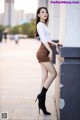 A woman in a white shirt and brown skirt leaning against a wall.