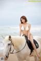 A woman in a white bikini riding a white horse on the beach.