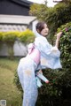 A woman in a blue and pink kimono leaning against a bush.