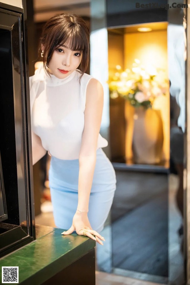 A woman in a white top and blue skirt leaning against a window.